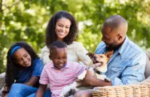 A joyful family of four with a small dog, sitting outdoors on a sunny day. The parents are smiling as they watch their daughter and son playfully interact with the energetic, panting dog.