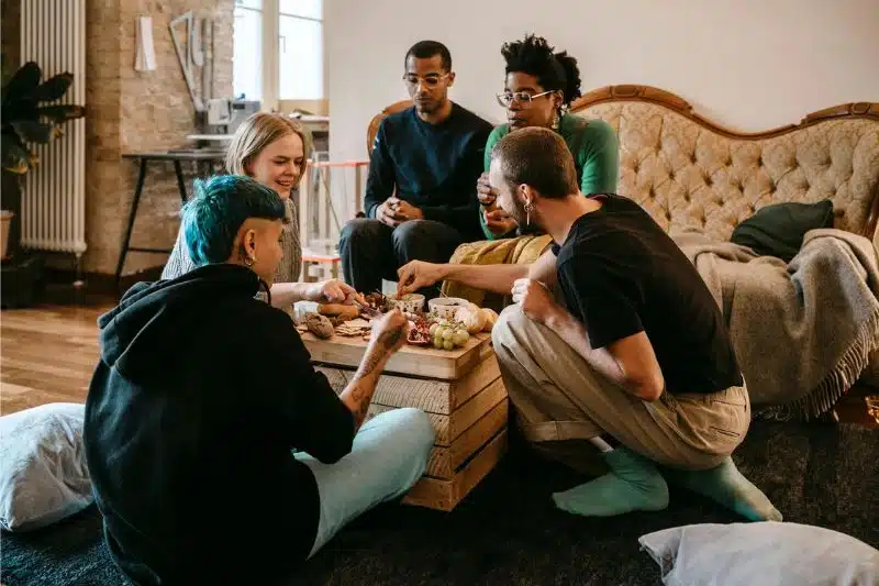 A group of five young people enjoying a casual get-together around a wooden table with snacks, engaged in a lively conversation in a cozy living room setting.